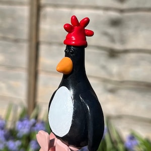 Può includere: Statuetta di pinguino fatta a mano con corpo nero, pancia bianca, becco arancione e cappello rosso. Il pinguino tiene in mano un oggetto rosa. La statuetta è ambientata su uno sfondo sfocato di vegetazione e una staccionata di legno.