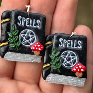 以下が含まれることがあります： 黒い本型のイヤリングのペアで、白い文字でSPELLSと書かれています。各イヤリングには、白い五芒星、緑の葉の小枝、白い斑点のある小さな赤いキノコが描かれています。本のページはシルバーのグリッター仕上げになっています。
