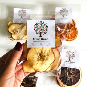 May include: Four clear plastic bags filled with dried fruit slices. The bags are labeled "Fresh Dried" with a logo of a tree with orange fruit. The fruit slices include apple, orange, and pear.