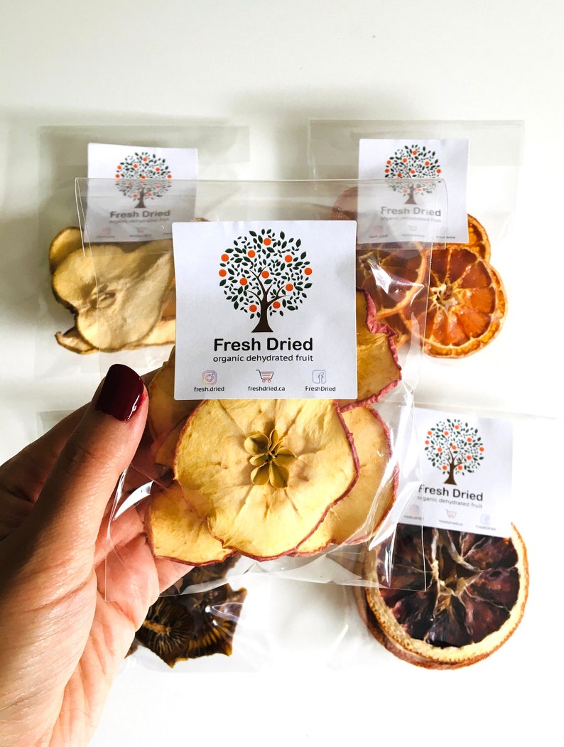 May include: Four clear plastic bags filled with dried fruit slices. The bags are labeled "Fresh Dried" with a logo of a tree with orange fruit. The fruit slices include apple, orange, and pear.