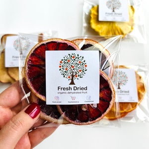 May include: A clear plastic bag filled with dried blood orange slices. The bag has a white label with the text "Fresh Dried" and "organic dehydrated fruit". The label also includes social media icons for Instagram, Facebook, and a shopping cart.