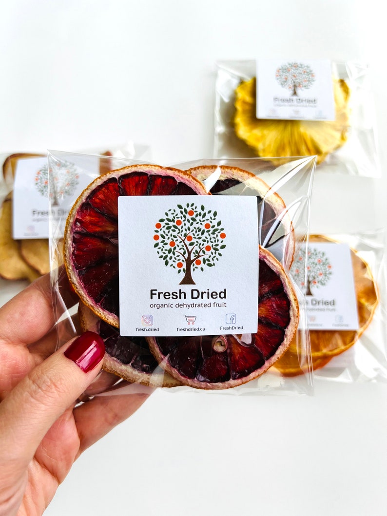 May include: A clear plastic bag filled with dried blood orange slices. The bag has a white label with the text "Fresh Dried" and "organic dehydrated fruit". The label also includes social media icons for Instagram, Facebook, and a shopping cart.