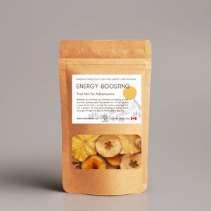 Op de afbeelding: Een bruine papieren zak met een doorzichtig venster dat gedroogd fruit laat zien. De zak is gelabeld met "ENERGY-BOOSTING Trail Mix for Adventurers". Het label vermeldt ook "ORGANIC DRIED FRUIT - NOTHING ADDED - 100% NATURAL".