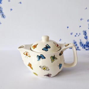 May include: Cream ceramic teapot with lid, decorated with colourful butterfly illustrations. The teapot has a curved spout and a handle. The butterflies are in various colours, including blue, orange, yellow, and pink. A charming addition to any kitchen.