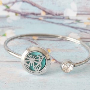 May include: Silver-coloured bangle bracelet with a round charm featuring a butterfly cutout and a turquoise backing. The bracelet also has a clear, faceted crystal accent. A stylish piece of jewellery.