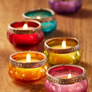 May include: A selection of lit votive candles in glass holders with silver rims. The candles are in various colours including red, orange, yellow, green, teal, pink, and purple, on a wooden surface.