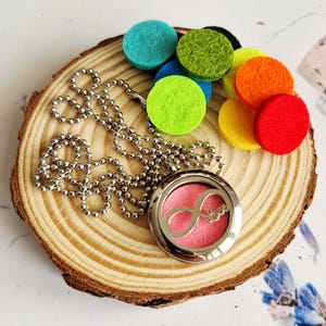 May include: A silver aromatherapy necklace with a pink interior and the word "Love" inside an infinity symbol. The necklace is on a wooden slice, with colourful felt pads in blue, green, yellow, orange, and red.