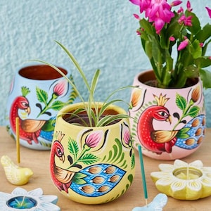 May include: Three hand-painted ceramic pots with peacock designs in blue, yellow, and pink, each holding plants. Decorative bird-shaped incense holders and flower-shaped candle holders are also present, creating a colourful and artistic display.