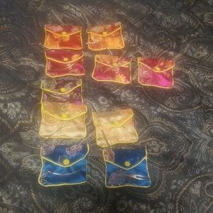 May include: Nine small, colorful, satin pouches with yellow trim and a gold zipper. The pouches are arranged in three rows on a dark patterned fabric.
