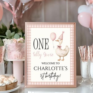 Könnte beinhalten: Ein gerahmtes Schild mit einem rosa Vichy-Karo-Rand zeigt den Text "ONE Silly Goose" und "Welcome to CHARLOTTE'S 1st birthday!" Eine Aquarellillustration einer Gans mit Partyhut und Fliege ist ebenfalls enthalten.
