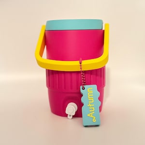 May include: A bright pink water cooler with a turquoise lid and a yellow handle. The cooler has a white spigot and a turquoise tag that says "Autumn" in yellow.