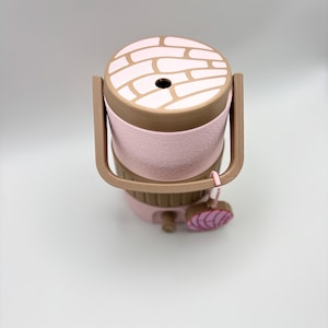 May include: A pink and brown insulated lunch box with a handle. The lid has a brick pattern design. A small pink charm hangs from the handle. The lunch box is designed to keep food at the desired temperature.