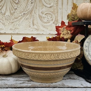 May include: A beige ceramic mixing bowl featuring a wide dark brown horizontal band, thin blue accent stripes, and a textured diamond pattern embossed around the upper and lower rim. The bowl has a rounded shape and a smooth interior finish.