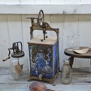 May include: Vintage blue and white metal butter churn with a hand crank and a wooden top. The churn is sitting on a wooden table with two glass bottles and a wooden spoon.