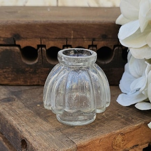 May include: A clear, vintage glass inkwell with a fluted, scalloped design. The small, decorative inkwell sits on a weathered wooden surface. The inkwell has a round opening and a bulbous body. The background includes a wooden box and white flowers.