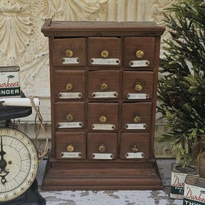 May include: A vintage wooden spice rack with twelve small drawers. Each drawer has a brass knob and a label. The rack is a dark brown color and sits on a small base. The labels are white with black text.
