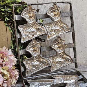 May include: Vintage metal mold for making chocolate lambs. The mold is made of silver-colored metal and has four lamb shapes. The mold is open and the lamb shapes are visible.