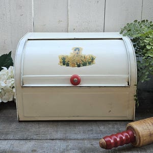 May include: Cream-colored vintage bread box with a roll-top lid and a red knob. The box features a decorative sticker with a floral design. A wooden rolling pin with a red handle rests nearby.