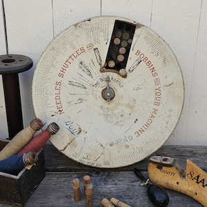 May include: An antique sewing machine parts display wheel with the text "Needles, Shuttles and Bobbins for Your Machine". The wheel is off-white with red lettering and a central dial. Various sewing notions are arranged around the wheel.