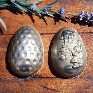 May include: Two vintage silver-colored metal egg molds. One mold has a dimpled surface, and the other features a relief design of children and a structure. The molds are on a wooden surface.