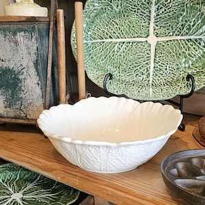 May include: A white ceramic bowl with a cabbage leaf design. The bowl has a scalloped edge and is sitting on a wooden shelf. A large green cabbage leaf plate is behind the bowl.