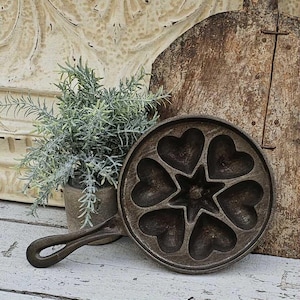 May include: A cast iron pan with heart-shaped molds and a star in the center. The pan has a long handle and is dark brown. A small potted plant with green foliage is to the left, and a wooden cutting board is in the background.