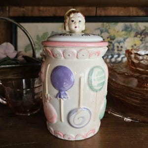 May include: A ceramic canister with a cream-colored base featuring embossed pastel-colored lollipops in purple, pink, and green. The lid is topped with a sculpted child's head and features a pink rim with scalloped detailing around the top edge.
