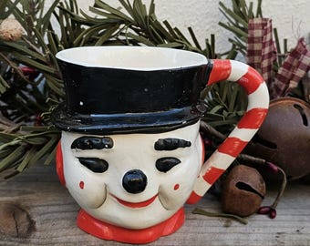 Puede incluir: Una taza de cerámica con forma de muñeco de nieve con un sombrero de copa negro y un asa a rayas rojas y blancas. La taza tiene una cara sonriente con ojos negros y una nariz negra. La base y las orejeras son rojas.