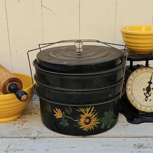 May include: A black, three-tiered vintage food carrier with a sunflower design. The carrier has a handle and a clear glass knob on the lid. A yellow bowl and a rolling pin are in the background.