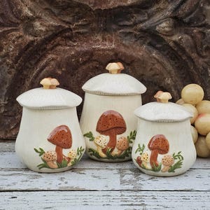 May include: Three off-white ceramic canisters with mushroom designs. Each canister has a lid with a small, decorative knob. The mushroom illustrations feature brown caps and white stems, with green foliage accents. The canisters vary in size.