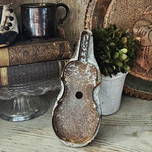 May include: A vintage-style violin-shaped cookie cutter with a distressed, rusty metal finish. The cutter has a white painted border and a small hole in the center. It is displayed on a wooden surface with other antique items.