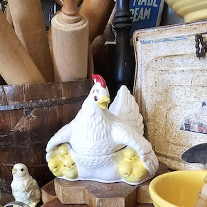 May include: A white ceramic hen figurine with a red comb and yellow beak, featuring small yellow chicks tucked under its wings. The hen is sculpted with textured feathers and sits in a nesting position, serving as a decorative lidded container.