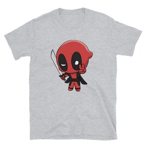 Puede incluir: Una camiseta gris con una imagen de dibujos animados de Deadpool sosteniendo una katana y haciendo un signo de paz. El personaje es rojo y negro con un ojo blanco.
