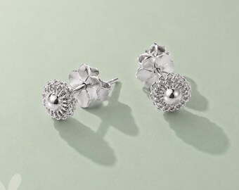 May include: A pair of silver stud earrings. Each earring features a central, smooth, spherical bead surrounded by a delicate, textured, floral-like design. The earring posts have a flower-shaped backing. The earrings are displayed on a light green surface.