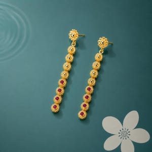 May include: A pair of gold-colored dangle earrings. Each earring features a small flower-shaped stud, a series of gold beads, and red gemstones. The earrings are displayed against a teal background.