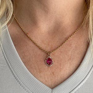 May include: A gold-colored necklace with a pendant featuring a pink gemstone. The pendant is set in a decorative frame. The necklace is worn around the neck, and the background is a light gray shirt.