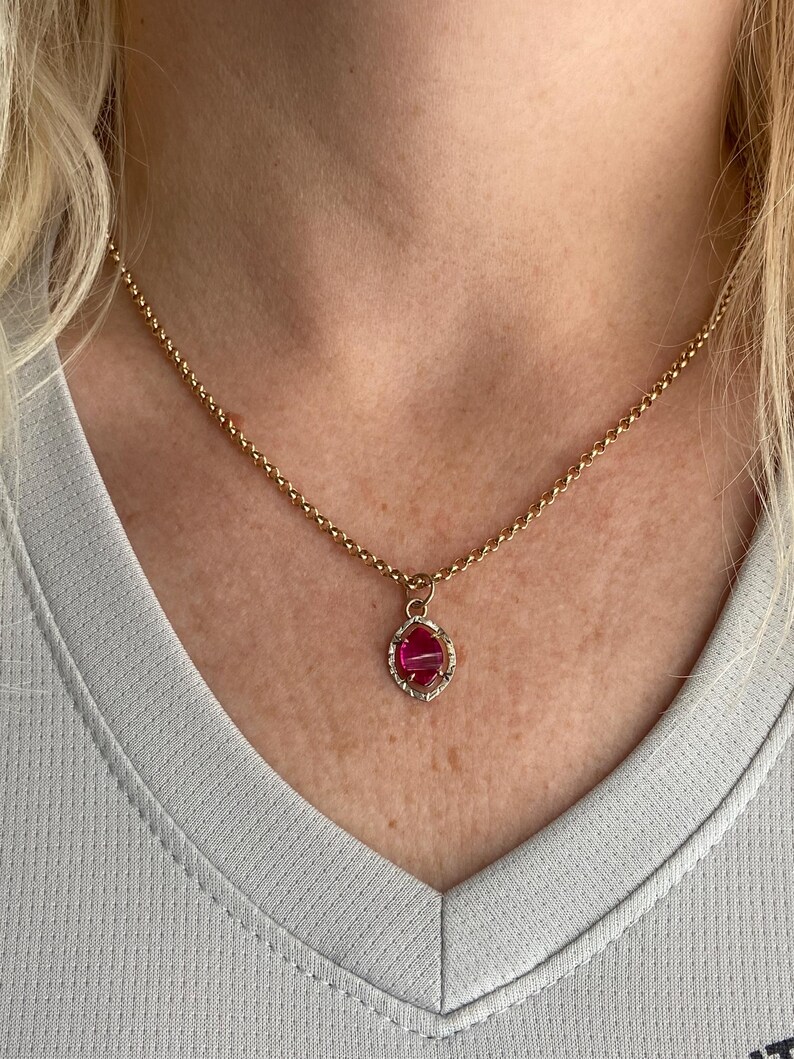 May include: A gold-colored necklace with a pendant featuring a pink gemstone. The pendant is set in a decorative frame. The necklace is worn around the neck, and the background is a light gray shirt.