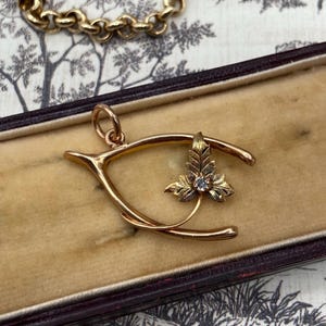 May include: A gold-colored wishbone pendant with a leaf and gemstone design. The pendant is displayed in a vintage jewelry box with a patterned interior. A gold chain is visible in the background.