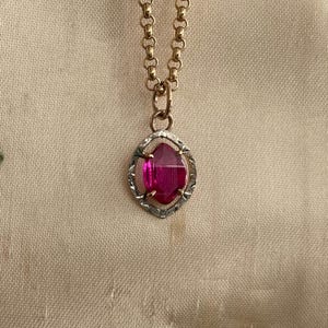 May include: A gold-toned chain necklace with a pendant featuring a vibrant pink gemstone. The gemstone is set in a silver-toned, geometric frame. The necklace is displayed on a neutral-colored fabric.