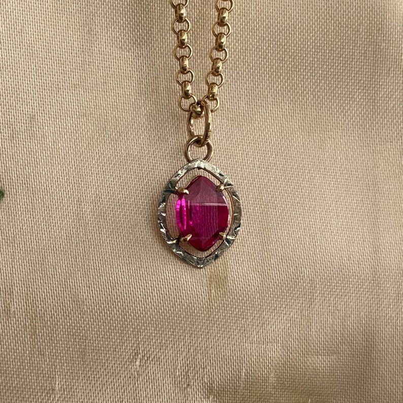 May include: A gold-toned chain necklace with a pendant featuring a vibrant pink gemstone. The gemstone is set in a silver-toned, geometric frame. The necklace is displayed on a neutral-colored fabric.