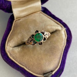 May include: A gold ring with a large, round, emerald-green gemstone at the center, surrounded by a floral design. The ring is set in a vintage-style setting and is displayed in a purple velvet ring box.