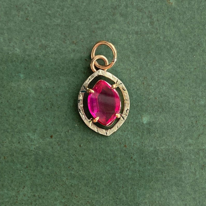 May include: A gold pendant with a marquise-cut, vibrant pink gemstone. The gemstone is set within a detailed, textured gold frame. The pendant has a small, looped bail for a chain.