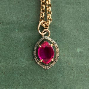 May include: A gold-toned necklace with a large, oval-shaped, magenta-colored gemstone pendant. The pendant is set in a decorative frame. The chain is a classic link style, and the background is a solid green.