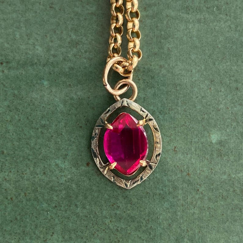 May include: A gold-toned necklace with a large, oval-shaped, magenta-colored gemstone pendant. The pendant is set in a decorative frame. The chain is a classic link style, and the background is a solid green.