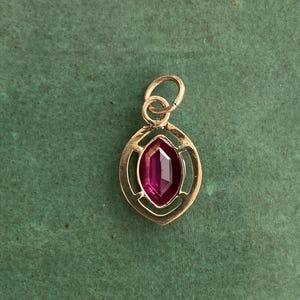 May include: A gold-colored pendant with a marquise-cut, deep red gemstone. The gemstone is set within a decorative, open-work frame. The pendant has a small loop at the top for a chain. The background is a solid, muted green.