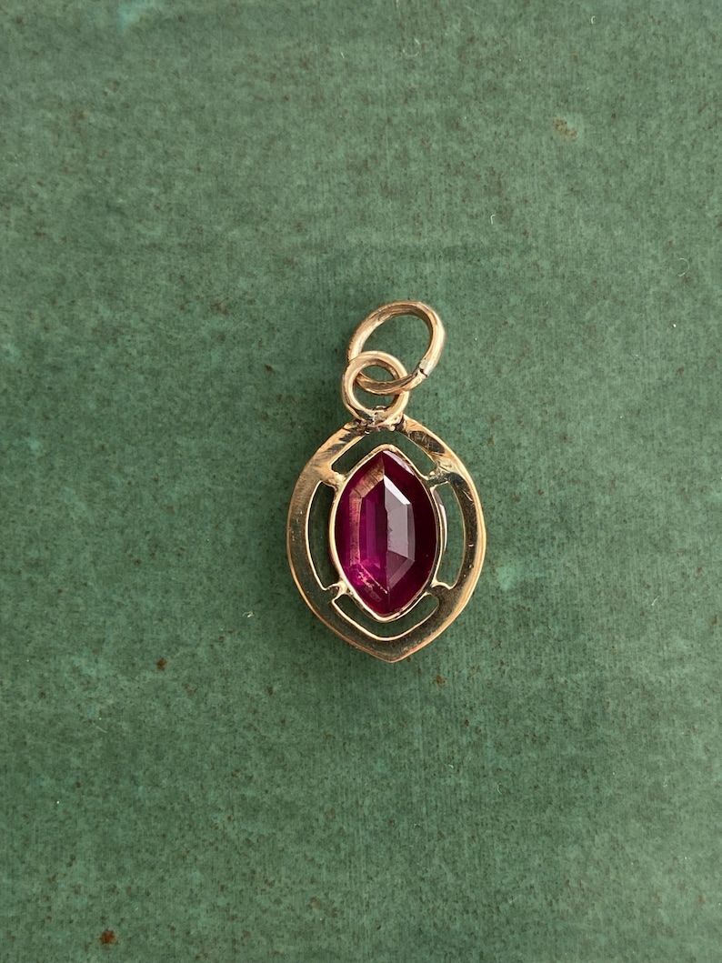 May include: A gold-colored pendant with a marquise-cut, deep red gemstone. The gemstone is set within a decorative, open-work frame. The pendant has a small loop at the top for a chain. The background is a solid, muted green.