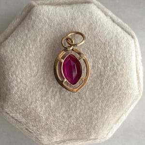 May include: A gold pendant with a marquise-cut, deep pink gemstone. The pendant has an openwork design with a loop at the top for a chain. The gemstone is set in a gold frame. The pendant is displayed on a light beige surface.