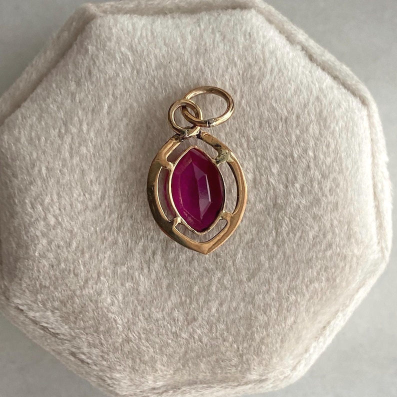 May include: A gold pendant with a marquise-cut, deep pink gemstone. The pendant has an openwork design with a loop at the top for a chain. The gemstone is set in a gold frame. The pendant is displayed on a light beige surface.