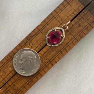 May include: A vintage pendant with a large, oval-shaped, faceted, ruby-red gemstone set in a gold-tone frame. The pendant is displayed on a wooden ruler next to a silver dime from 1965.