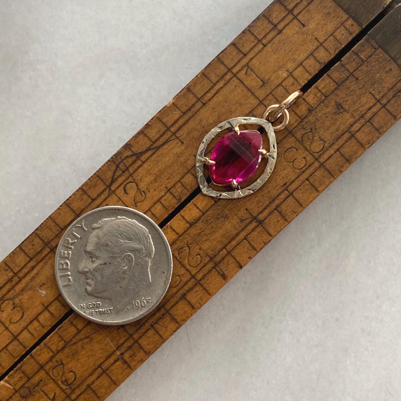 May include: A vintage pendant with a large, oval-shaped, faceted, ruby-red gemstone set in a gold-tone frame. The pendant is displayed on a wooden ruler next to a silver dime from 1965.